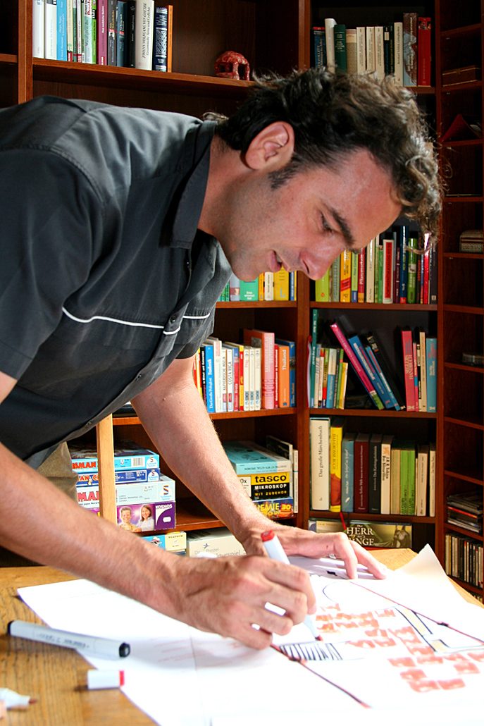 Bernd Bartelmeß zeichnet am Schreibtisch vor dem Bücherregal